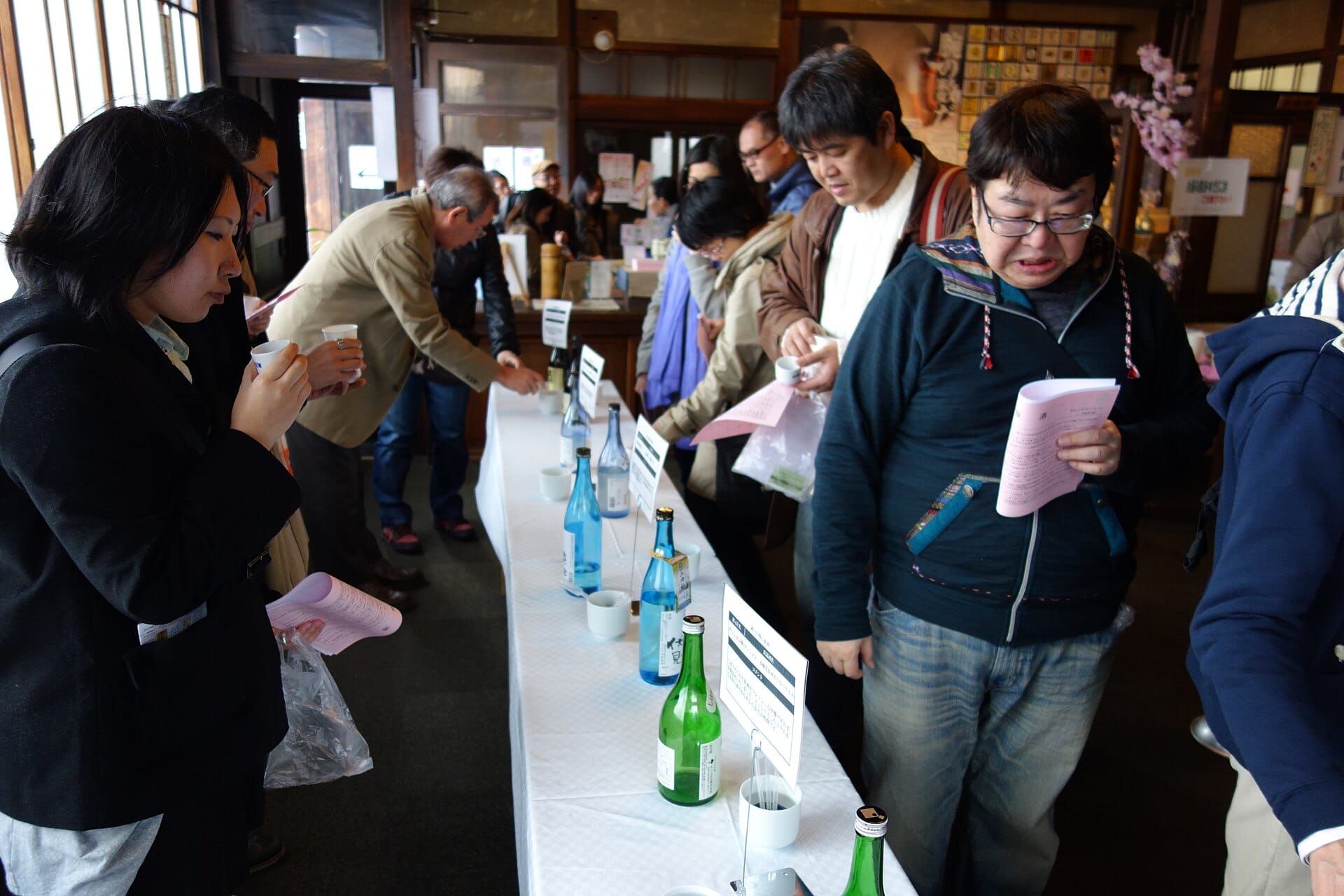 Adults holding small cups and printed tasting sheets beside a table lined with sake bottles at a crowded indoor tasting.